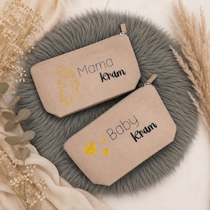 Könnte beinhalten: Zwei beige Reißverschlusstaschen mit den Aufschriften "Mama Kram" und "Baby Kram". Die "Mama" Tasche hat ein goldenes Glitzerdesign, die "Baby" Tasche goldene Glitzerherzen.