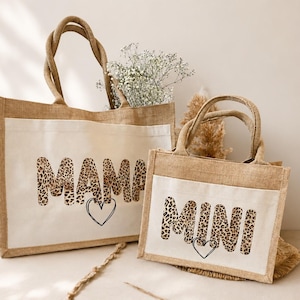 K&ouml;nnte beinhalten: Zwei rechteckige Jute-Tragetaschen mit wei&szlig;en Canvas-Frontpaneelen. Die gr&ouml;&szlig;ere Tasche zeigt das Wort MAMA in einem Leopardenmuster mit einem schwarzen Herzumriss darunter. Die kleinere Tasche zeigt das Wort MINI im gleichen Leopardenstil.