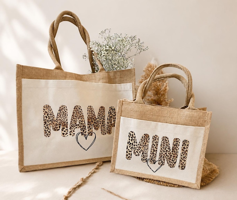 K&ouml;nnte beinhalten: Zwei rechteckige Jute-Tragetaschen mit wei&szlig;en Canvas-Frontpaneelen. Die gr&ouml;&szlig;ere Tasche zeigt das Wort MAMA in einem Leopardenmuster mit einem schwarzen Herzumriss darunter. Die kleinere Tasche zeigt das Wort MINI im gleichen Leopardenstil.