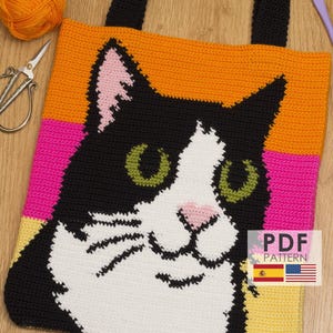 Puede incluir: Bolso tote tejido a crochet con diseño de cara de gato. El bolso tiene un asa negra y una cara de gato colorida con detalles en naranja, rosa, negro, blanco y verde. Se ven hilos, tijeras y un ganchillo.