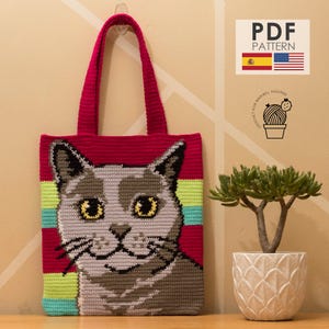Puede incluir: Un bolso tote de crochet hecho a mano con un diseño de cara de gato. El bolso tiene un fondo rojo con una cara de gato en tonos grises, amarillos y negros. Las asas del bolso también son rojas. El diseño incluye el texto "PDF PATTERN".