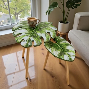 May include: Two small wooden tables with a monstera leaf design. The tables have a green and white painted leaf top and three wooden legs. A cup of coffee sits on the larger table. The tables are on a hardwood floor.