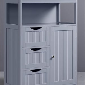 May include: A gray wooden bathroom cabinet with three drawers and a cabinet door. The cabinet has a top shelf and an open side shelf. The drawers and door have vertical paneling and curved handles. The cabinet is painted in a solid gray color.