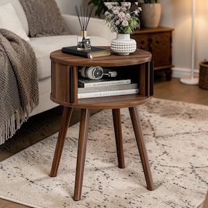 May include: A round, dark brown wooden side table with three angled legs. The table has a storage compartment with books and headphones. A vase of flowers, a book, and a fragrance diffuser sit on top. The table is on a patterned rug.