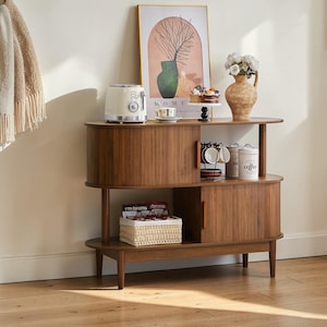 May include: A mid-century modern style wooden cabinet with three shelves. The top shelf holds a cream-colored toaster, a framed art print, and a vase with flowers. The middle shelf has coffee cups and canisters. The bottom shelf has a woven basket.