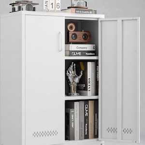 May include: White metal cabinet with open door revealing books and decorative items. A small gramophone, a wooden speaker, and a metal hand sculpture are visible. The cabinet has a simple design with a handle and ventilation holes.