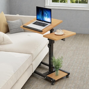 May include: A two-tiered, adjustable laptop desk with a light wood finish. The top tier holds a laptop, while the lower tier has a teacup and saucer. The desk has a black metal frame and sits on wheels.