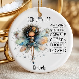 May include: A white ceramic Christmas ornament with a gold string. The ornament features a watercolor illustration of a fairy with text that reads "GOD SAYS I AM AMAZING, BEAUTIFUL, CAPABLE, CHOSEN, ENOUGH, LOVED" and the name "Kimberly".