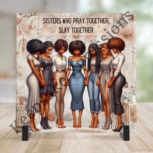 May include: A square decorative print featuring seven women of color holding hands, with the text "SISTERS WHO PRAY TOGETHER, SLAY TOGETHER" at the top. The women are dressed in various outfits, including dresses, jeans, and blouses, in shades of gray, blue, and beige.