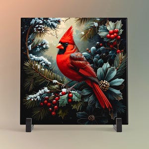 May include: A square decorative tile featuring a vibrant red cardinal perched on a branch. The bird is surrounded by winter foliage, including snow-covered pine needles, holly berries, and blue-toned flowers, set against a dark background. The tile is displayed on a black stand.