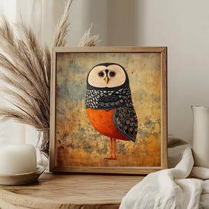 May include: Framed art print featuring a stylized owl illustration. The owl has a patterned black and white head, a vibrant orange body, and long orange legs. The artwork is set against a textured, muted background, and framed in a light wood.