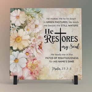 May include: A square decorative plaque featuring floral artwork in soft pinks, whites, and yellows. The plaque includes the text "He Restores my Soul" and a Bible verse. The plaque is displayed on a black stand.