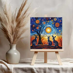 May include: A painting on canvas depicting three kangaroos standing in front of a tree with a bright yellow sun and a blue starry sky. The painting is on a wooden easel.