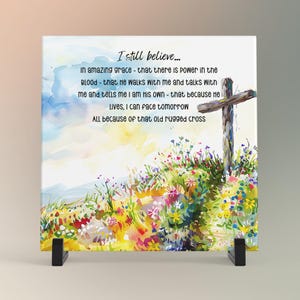 May include: A square canvas print featuring a watercolor painting of a cross and a field of wildflowers. The text "I still believe..." and other inspirational phrases are written on the canvas. The artwork is displayed on a black stand.