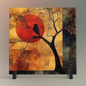 May include: Square art print featuring a black bird perched on a tree branch against a large red sun. The background has a textured, golden appearance with a dark tree silhouette. The artwork is displayed on a black stand.