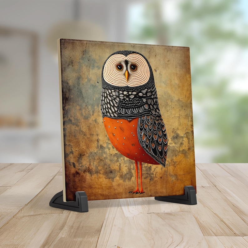 May include: A decorative art piece featuring a stylized owl illustration. The owl has a white and black patterned head, orange body, and brown wings. The artwork is displayed on a rectangular panel supported by a black stand.