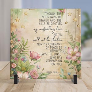 May include: A square decorative plaque with a floral and botanical design, featuring a quote from Isaiah 54:10. The artwork includes pink and white flowers, green leaves, and gold stars, set against a neutral background. The plaque is displayed on a black stand.