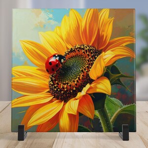 May include: A square canvas print featuring a vibrant sunflower with bright yellow petals and a dark center. A red ladybug with black spots rests on the flower. The artwork is displayed on a black stand.