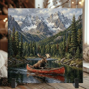 May include: A square decorative tile featuring a detailed illustration of a brown moose paddling a red canoe on a calm lake. The background displays a dense green pine forest and towering, snow-capped mountain peaks under a cloudy sky.