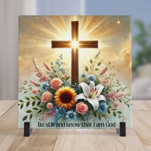 May include: A square decorative piece featuring a wooden cross with a bright light emanating from behind it. The cross is surrounded by a floral arrangement of roses, sunflowers, and lilies. The text "Be still and know that I am God" is at the bottom.