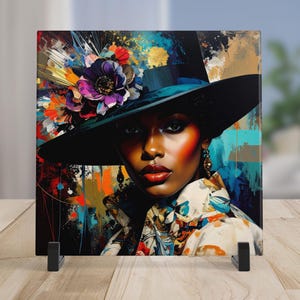 May include: Square artwork featuring a portrait of a person wearing a black hat adorned with colorful flowers. The artwork has a vibrant, abstract background with splashes of color and gold accents. The person has dark skin, red lips, and is wearing a patterned shirt.