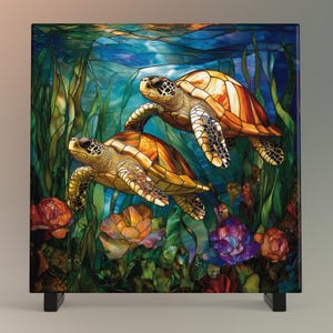 May include: A square decorative panel featuring a vibrant underwater scene. Two sea turtles swim amidst colorful coral, seaweed, and flowers. The artwork is rendered in shades of blue, green, orange, and yellow, mounted on a black stand.