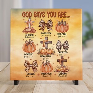 May include: Square canvas art with the text "GOD SAYS YOU ARE..." and various words like "CHOSEN" and "FORGIVEN". The artwork features pumpkins, bows, and crosses with fall colors. The canvas is supported by a black stand.