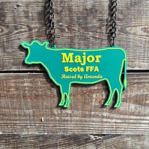 May include: A green cow-shaped sign with the text "Major Scots FFA" and "Raised by Amanda" in yellow lettering. The sign is hanging from a black chain against a wooden background.