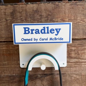 May include: A white rectangular wall-mounted bridle rack featuring a blue border and blue text that reads Bradley and Owned by Carol McBride. The rack includes a curved support piece for hanging equipment and is secured with visible metal screws.