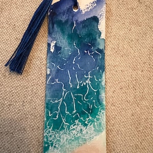 May include: A rectangular bookmark featuring a watercolor painting of an ocean scene with shades of blue, teal, and white foam. The design includes white line art depicting waves and a sandy beach at the bottom. A dark blue tassel is attached to the top.