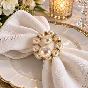 May include: A circular napkin ring featuring a cluster of white faux pearls and small gold-toned metal accents with embedded clear crystals. The ring holds a folded white linen napkin with a decorative hemstitched border detail.