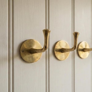 May include: Four brass coat hooks fixed to a white panelled wall. Each hook features a circular base and a curved, upward-facing hook. The hooks are evenly spaced, offering a simple, elegant design for hanging items.