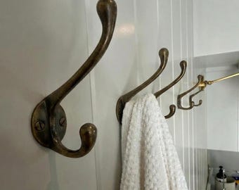 Puede incluir: Un juego de tres ganchos de pared de metal en tono latón antiguo con diseño de doble punta, con extremos redondeados arriba y abajo. Una toalla blanca texturizada cuelga del gancho central, junto a una lámpara dorada.