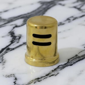 May include: A cylindrical, brass-colored object with two horizontal, black, rectangular cutouts. The object has a rounded top and a flared base. The background is a white and gray marble surface.