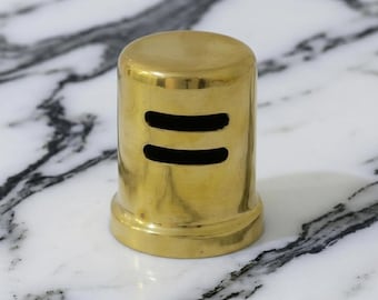 May include: A cylindrical, brass-colored object with two horizontal, black, rectangular cutouts. The object has a rounded top and a flared base. The background is a white and gray marble surface.