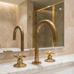 May include: Bathroom tap set with a brushed brass finish. The set features a high-arc spout and two cross-handle style knobs. The tap is mounted on a white marble countertop, with a mirror and shower visible in the background.