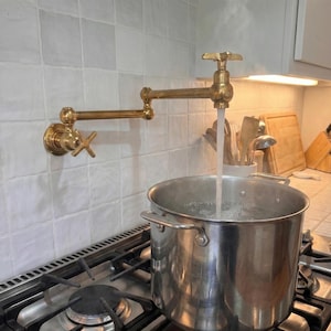 May include: A brass pot filler faucet is mounted on a white tiled wall. Water flows into a stainless steel pot on a gas stove. The faucet has a cross-shaped handle and an articulated arm.