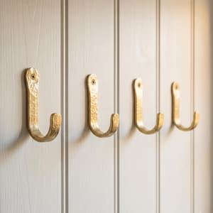 Könnte beinhalten: Vier goldfarbene Wandhaken aus Metall mit gehämmertem Finish. Jeder Haken hat eine geschwungene J-Form und ist mit einer einzelnen sichtbaren Schraube an der Oberseite befestigt. Die Haken sind in einer horizontalen Reihe an einer weißen Wand angebracht.