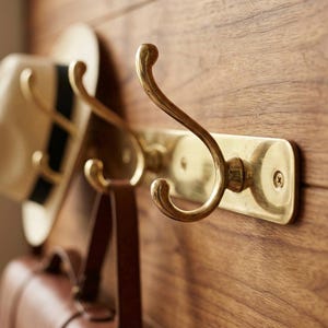 May include: A brass coat rack with three curved hooks, mounted on a wooden wall. A beige hat with a black stripe and a brown leather bag hang from the hooks. The rack is made of brass.