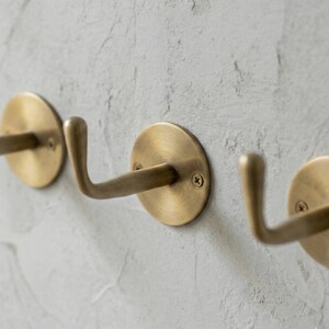 May include: Close-up of three brass wall hooks mounted on a textured, light gray wall. Each hook has a circular base and a curved arm, secured with screws. The hooks are designed for hanging items.