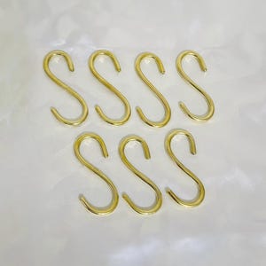May include: Seven gold-colored S-shaped hooks arranged on a white surface. The hooks are made of a shiny metal and have a simple, curved design. They are evenly spaced and appear to be new.
