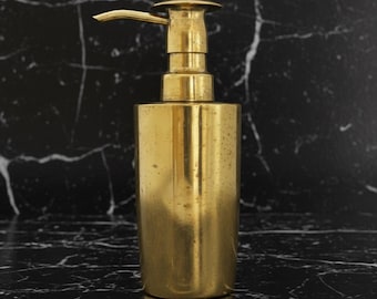 May include: A gold-colored soap dispenser with a pump top. The dispenser has a cylindrical body and a curved spout. The surface has a slightly aged appearance. The background is a black marble.