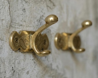 May include: Two antique brass wall hooks. Each hook has a decorative, ornate backplate with a curved hook. The hooks are mounted on a textured, light gray wall. The hooks are made of brass and have a shiny finish.