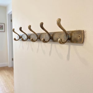 Può includere: Un attaccapanni da parete color ottone con cinque ganci curvi. I ganci sono montati su una piastra posteriore rettangolare con teste di viti visibili. L'attaccapanni è installato su una parete di colore chiaro, ideale per un corridoio o un ingresso.