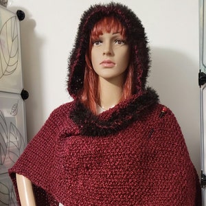 Puede incluir: Una capa con capucha burdeos con un diseño de punto texturizado. La capucha y el ribete presentan piel sintética negra. La capa se exhibe en un maniquí, mostrando su silueta drapeada y su estilo acogedor.