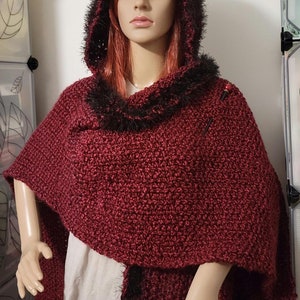 Pu&ograve; includere: Un mantello con cappuccio all'uncinetto bordeaux con finiture in finta pelliccia nera. Il mantello presenta una cucitura strutturata ed &egrave; esposto su un manichino. Il cappuccio e lo scollo sono rifiniti con finta pelliccia nera. Due spille decorative fissano il mantello.