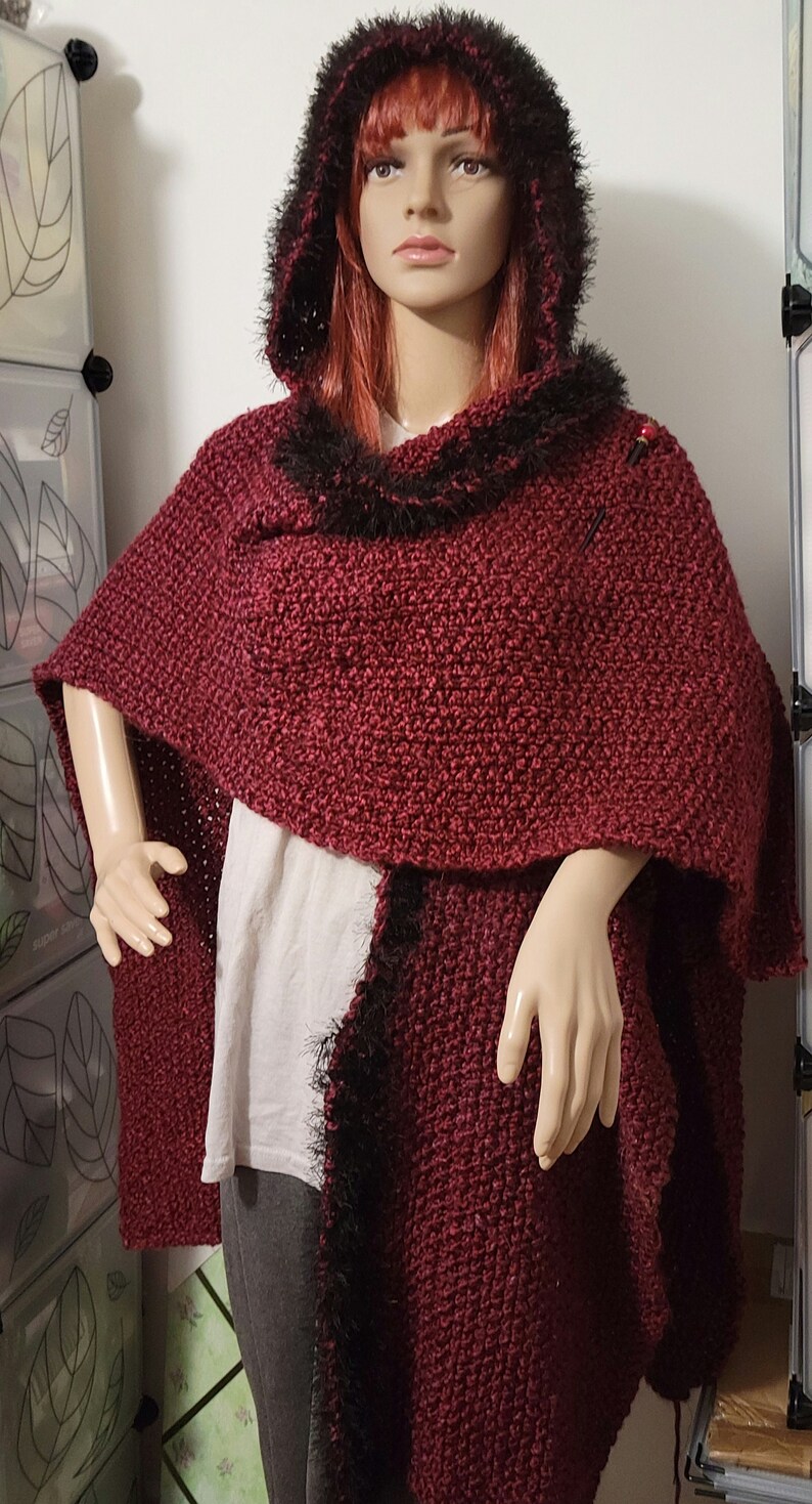 Pu&ograve; includere: Un mantello con cappuccio bordeaux con un design a maglia strutturata. Il cappuccio e l'orlo presentano ecopelliccia nera. Il mantello &egrave; esposto su un manichino, mostrando la sua silhouette drappeggiata e il suo stile accogliente.