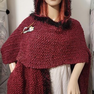 Pu&ograve; includere: Un mantello con cappuccio bordeaux con finiture in finta pelliccia nera. Il mantello &egrave; lavorato all'uncinetto e presenta una spilla decorativa. Il manichino indossa una camicia bianca sotto. Un accessorio comodo ed elegante.