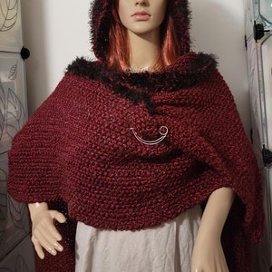 Pu&ograve; includere: Un mantello con cappuccio bordeaux con finiture in finta pelliccia nera. Il mantello &egrave; fissato con una spilla color argento. Il mantello &egrave; esposto su un manichino. Il mantello &egrave; realizzato in un tessuto a maglia spesso e strutturato.