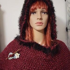 Pu&ograve; includere: Un poncho con cappuccio bordeaux con finiture nere e rosse. Il cappuccio &egrave; foderato in ecopelliccia nera. Una spilla di legno con un disegno di gatto &egrave; appuntata sul davanti. Il poncho &egrave; esposto su un manichino.